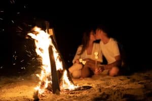 A young couple on the sea beach sits by the fire and toasts marshmallows on a stick. Romantic date by the fire. Marshmallow barbecue. Tourism and travel concept soft focus. copy space.